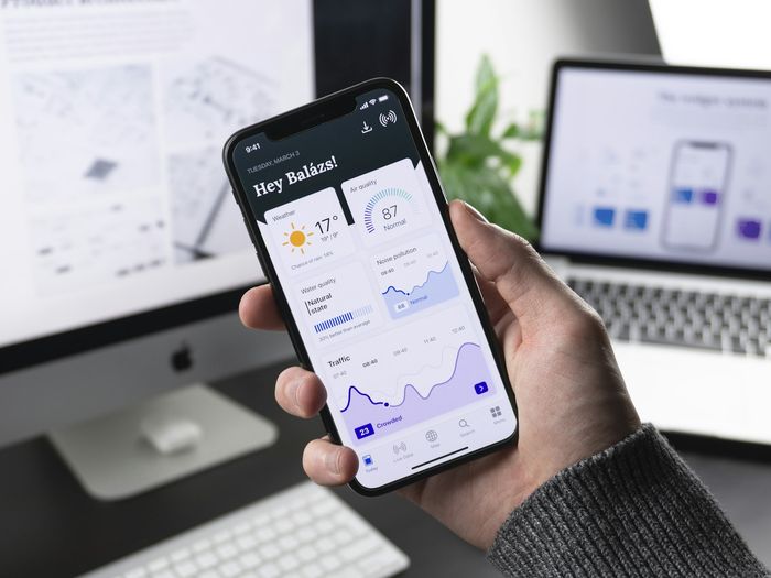 Pessoa segurando um celular com o painel de um aplicativo aberto, ao lado de um monitor com o mesmo sistema na versão web